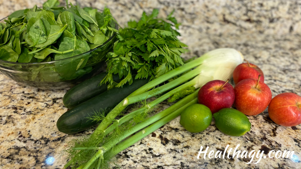 clear glass bowl of green spinach, 2 green cucumber, 1 bunch parsley, 1 green fennel bulb, 4 red apples 2 limes on a kitchen countertop