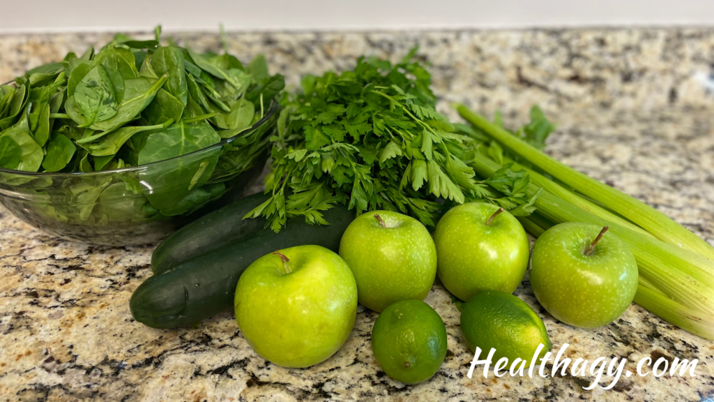 green spinach in a bowl, 2 green cucumbers, 1 bunch of parsley, 4 green apples, 1 bunch of celery, 2 green limes on a kitchen countertop